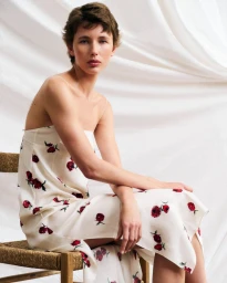 Model Nora Svenson seated on a woven wooden chair wearing a white floral slip dress in front of draped white fabric.