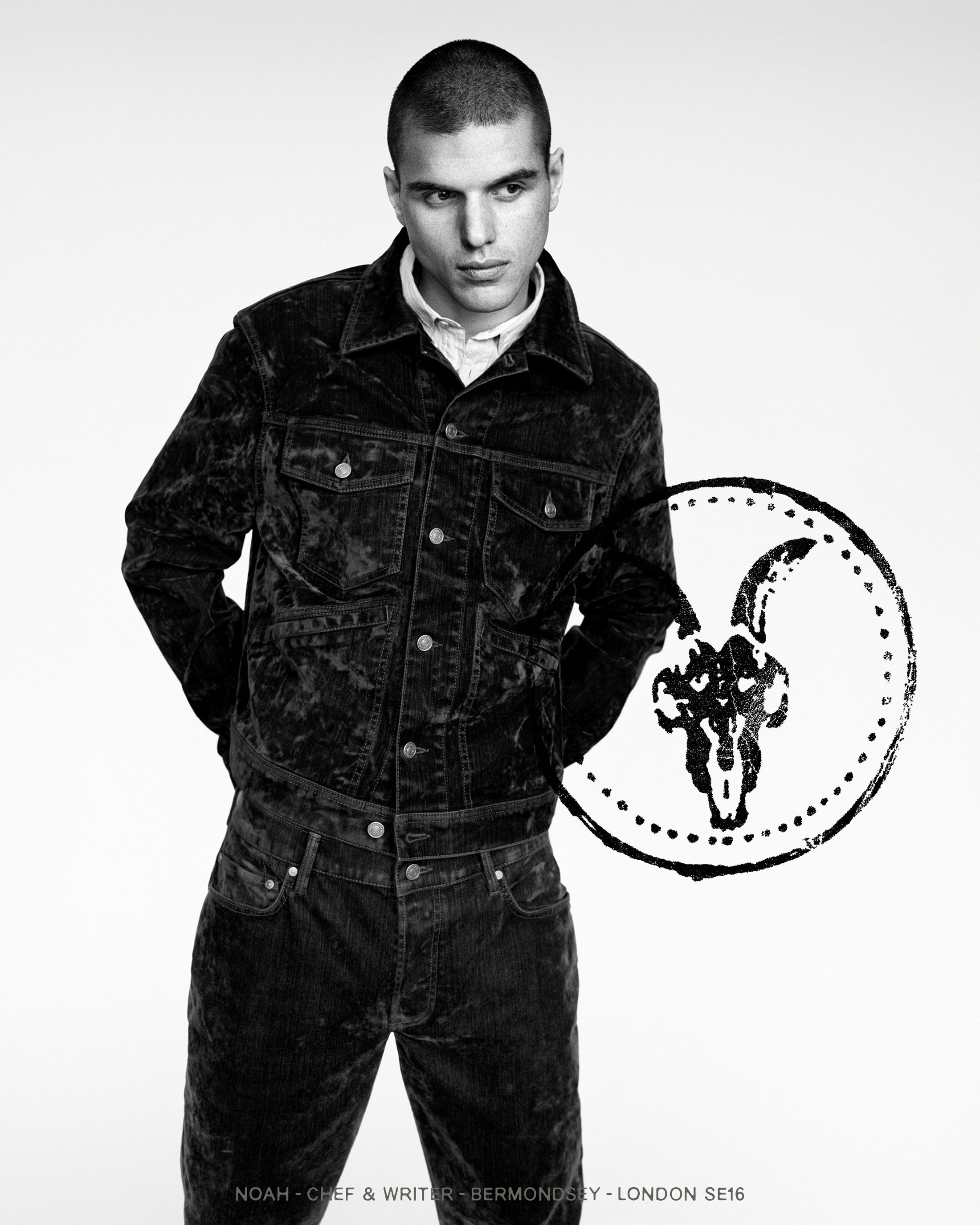 Black and white studio portrait of a man with a buzz cut wearing a dark textured jacket and matching trousers against a white background.
