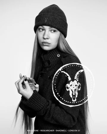Black and white portrait of a woman wearing a dark knit beanie and high-neck sweater with a ramskull logo overlay.