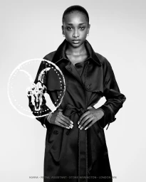 Black and white studio portrait of a Black woman with slicked hair wearing a belted dark satin trench coat.