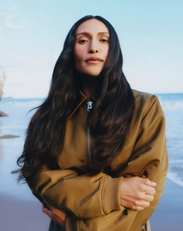 Woman with long dark hair wearing a khaki zip-up jacket posing with crossed arms on a beach with ocean waves.