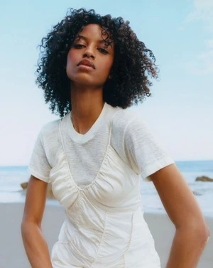 Model with curly dark hair wearing a layered white ruched dress over a t-shirt on a beach with ocean behind.
