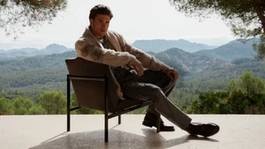 Model Max Knott seated in a modern armchair on a terrace wearing a knit cardigan and trousers against a scenic mountain backdrop.