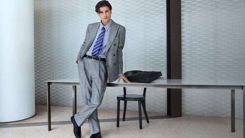 Model Max Knott wearing a grey double-breasted suit and striped blue tie leaning against an office desk for Mytheresa.