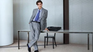 Model Max Knott wearing a grey double-breasted suit and striped blue tie leaning against an office desk for Mytheresa.