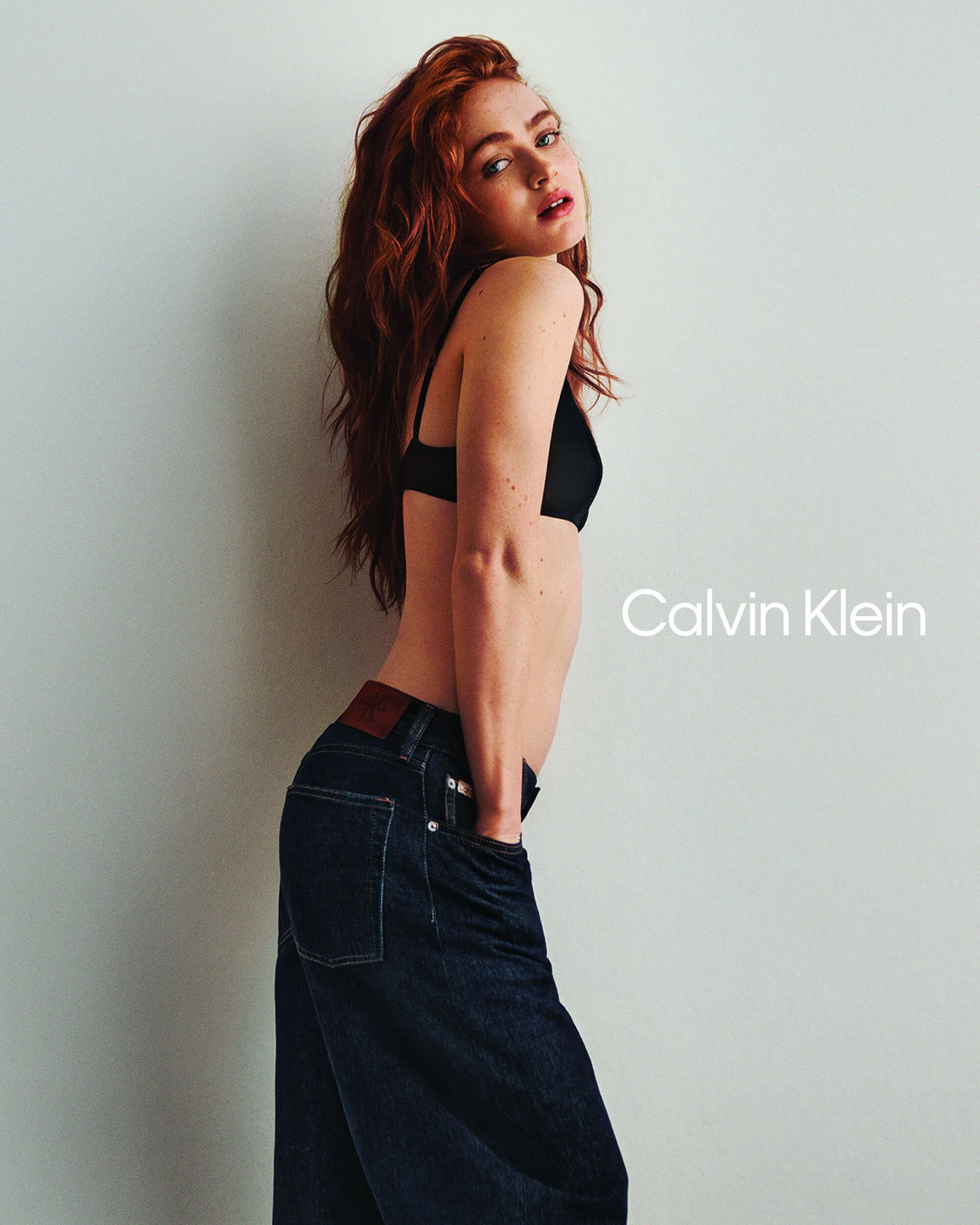 Sadie Sink wearing dark denim jeans and a black bra posing against a neutral studio backdrop for Calvin Klein.