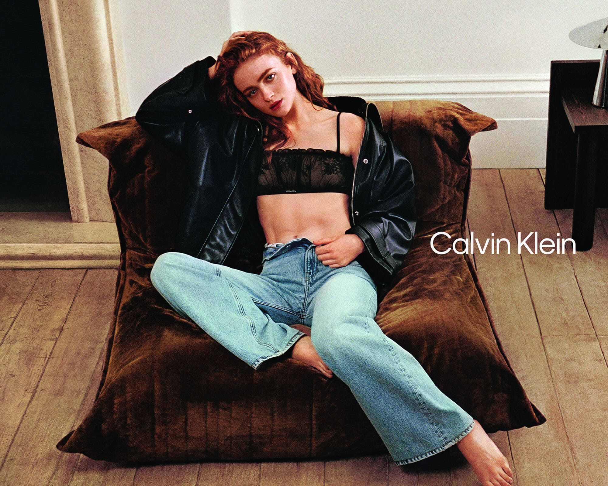 Sadie Sink sitting on a brown velvet floor cushion wearing light-wash denim jeans, a black lace bralette, and a black leather jacket.