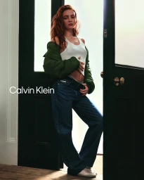 Sadie Sink wearing a white tank top, green jacket, and blue jeans standing in a dark doorway for Calvin Klein.
