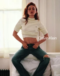 Sadie Sink sitting on a bed in a cream cropped ribbed sweater and ripped jeans for Calvin Klein.