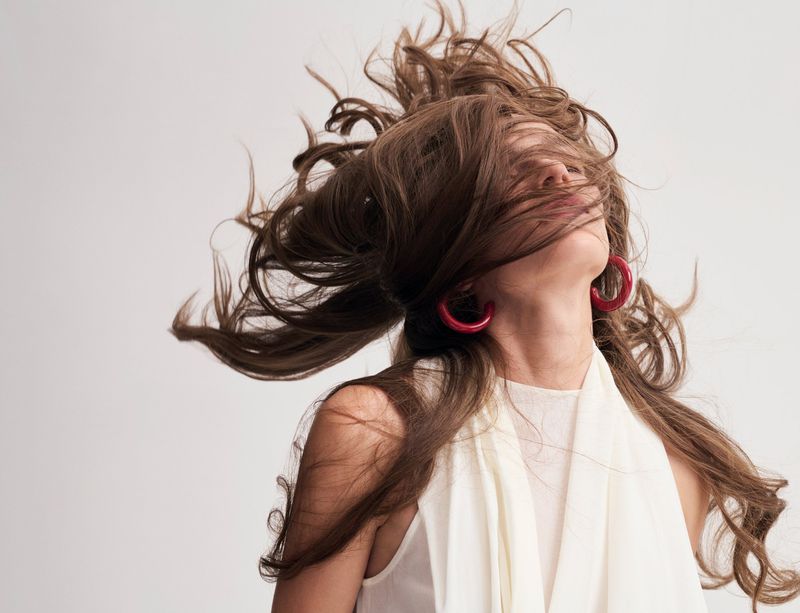 A woman tossing long brown hair across her face, wearing chunky red hoop earrings and a draped white top on a seamless backdrop.