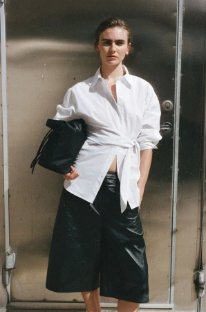 Model Mia Armstrong wearing a white wrap button-down shirt and black leather culottes against a brushed metal industrial door.