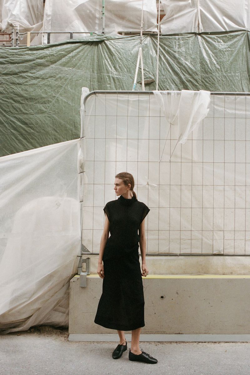 Model Mia Armstrong wearing a black sleeveless dress and flat shoes standing in front of construction scaffolding and plastic tarps.