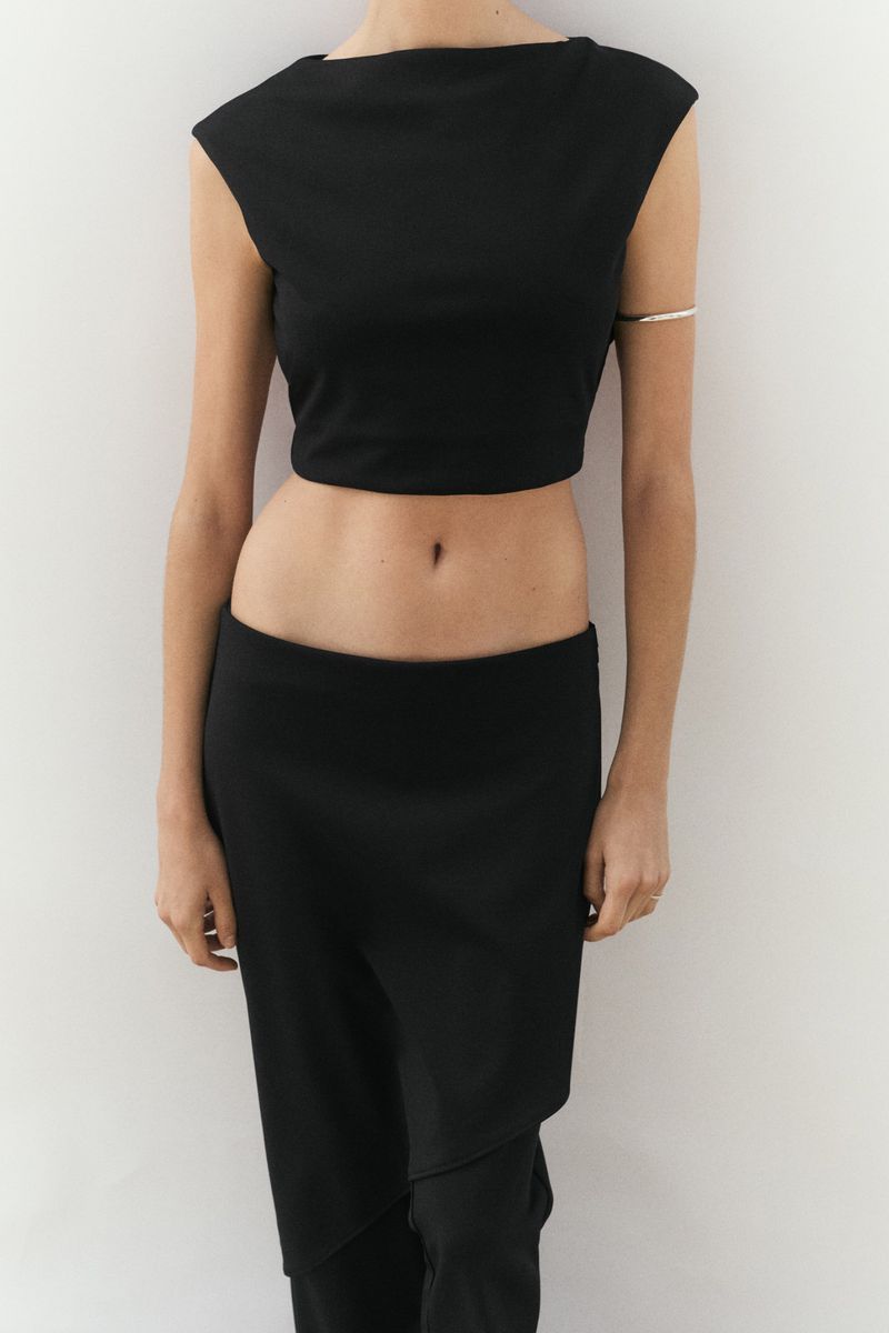 Model wearing a sleeveless black boatneck crop top, low-rise black trousers, and a thin silver arm cuff against a white studio background.