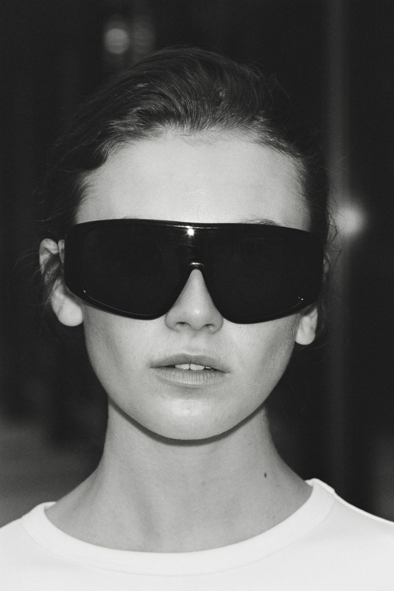 Model Mia Armstrong wearing oversized black shield sunglasses and a white crewneck shirt in a monochrome close-up portrait.