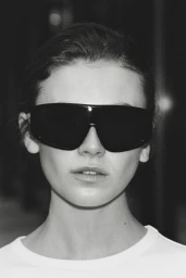 Model Mia Armstrong wearing oversized black shield sunglasses and a white crewneck shirt in a monochrome close-up portrait.