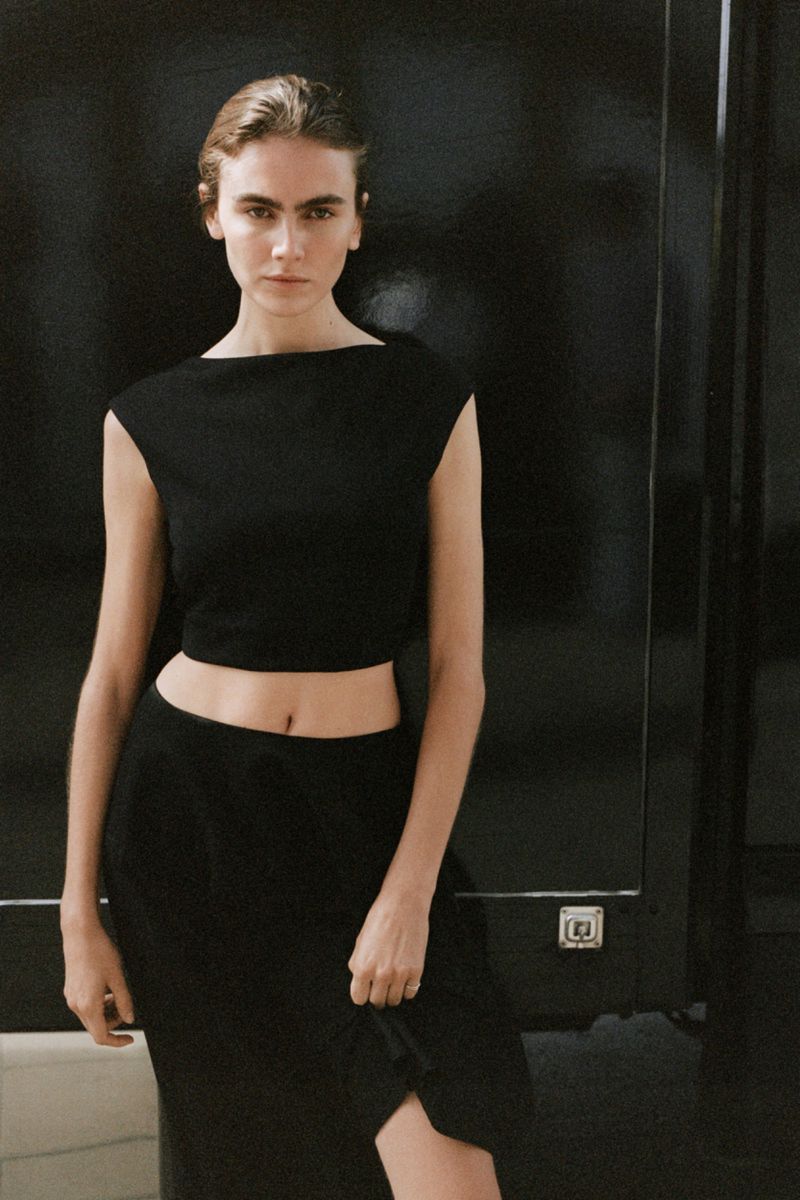 Model Mia Armstrong wearing a black sleeveless crop top and matching black skirt against a dark background for H&M.