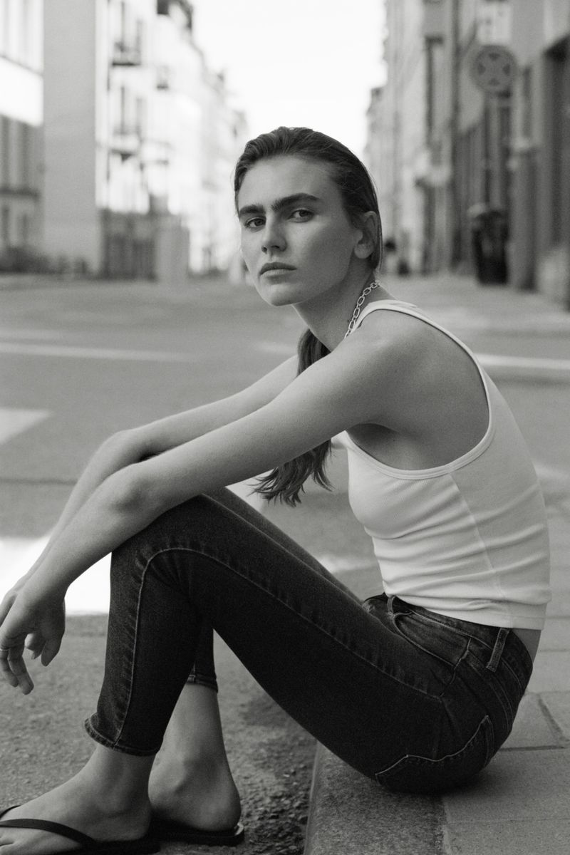 Model Mia Armstrong sitting on a street curb wearing a white tank top and dark jeans in a black and white fashion portrait.