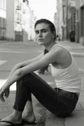 Model Mia Armstrong sitting on a street curb wearing a white tank top and dark jeans in a black and white fashion portrait.