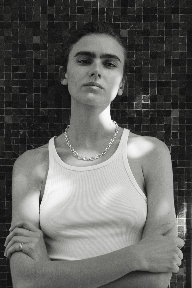 Black and white portrait of a woman with crossed arms wearing a white ribbed tank top and silver chain in front of a dark tile wall.