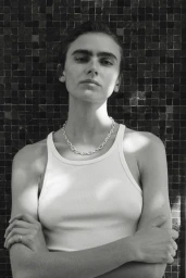 Black and white portrait of a woman with crossed arms wearing a white ribbed tank top and silver chain in front of a dark tile wall.