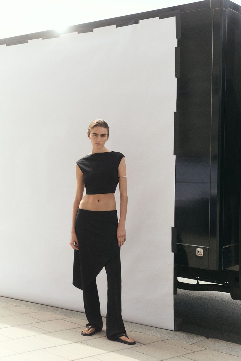 Model Mia Armstrong wearing a black sleeveless crop top and matching trousers in front of a white outdoor backdrop and truck.