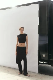 Model Mia Armstrong wearing a black sleeveless crop top and matching trousers in front of a white outdoor backdrop and truck.