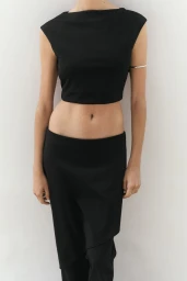 Model Mia Armstrong wearing a black sleeveless crop top and matching low-rise black pants against a white background.