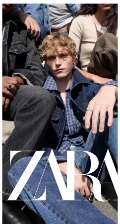 Blond male model in a denim jacket, blue plaid shirt, and jeans seated outdoors with others under bright sunlight.