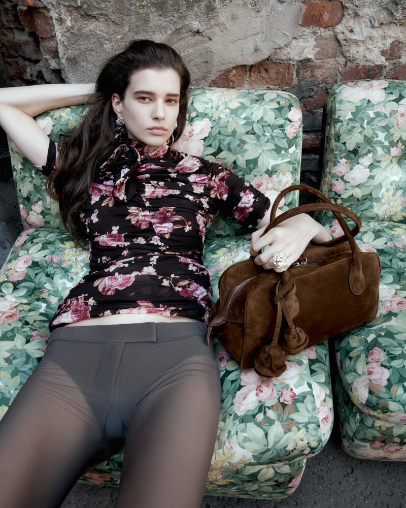 Woman lounges on a floral sofa with a brown handbag, wearing a floral top and grey tights, set against a textured grey wall.