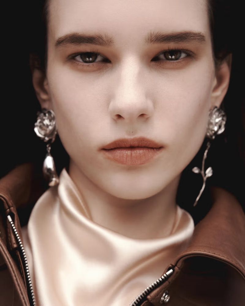 Close-up portrait of model Sandra Murray wearing sculptural silver rose earrings, a champagne satin scarf, and a brown leather jacket.