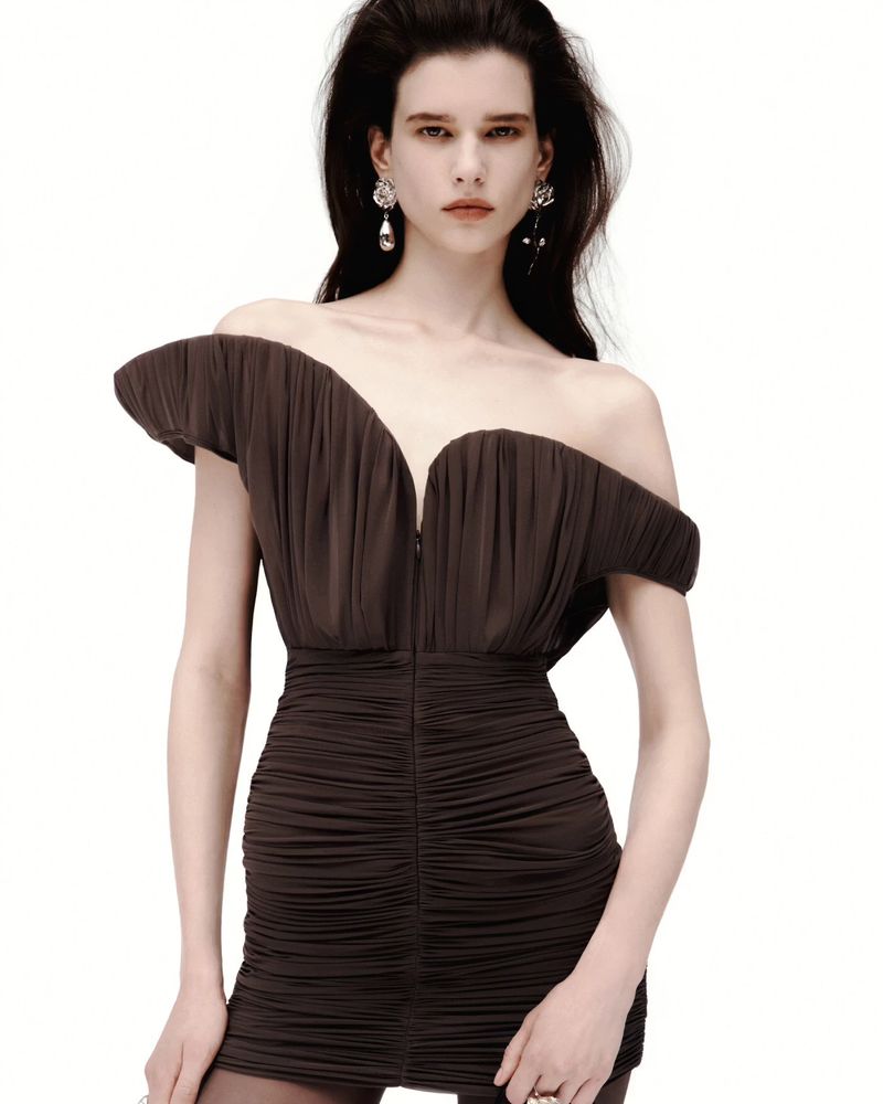 Elegant woman in a short brown dress with shoulder ruffles, dangling earrings, and long dark hair against a bright white backdrop.