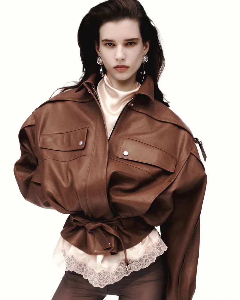 Fashion portrait of a woman wearing a brown leather jacket with a tied waist, a white turtleneck, and large earrings in a studio setting.