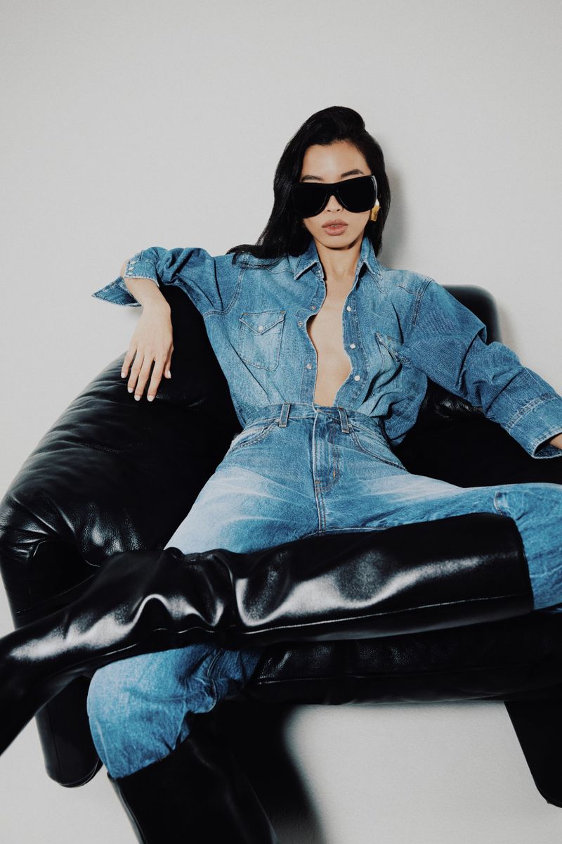 Model wearing oversized black sunglasses and double denim reclining in a black leather armchair against a plain white backdrop.