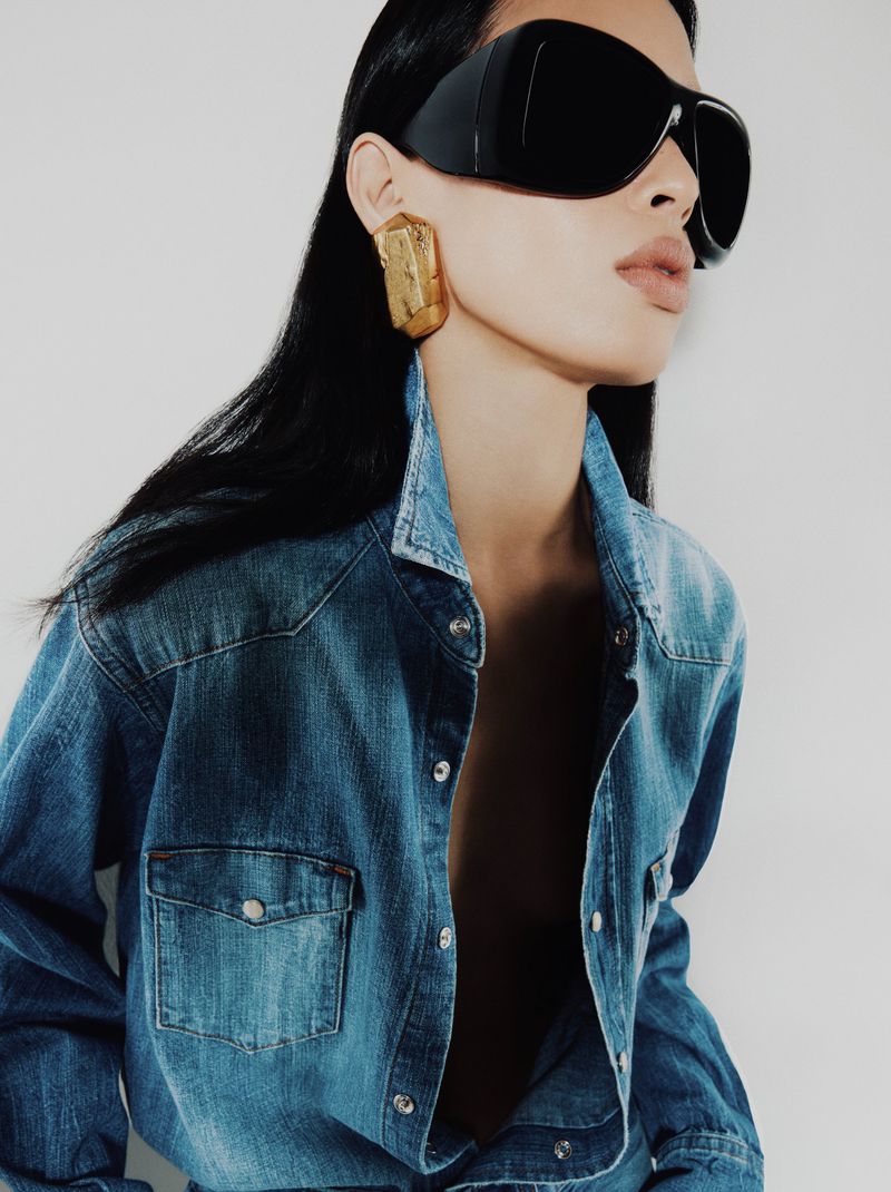 Model wearing oversized black shield sunglasses, a gold statement earring, and an unbuttoned blue denim shirt.