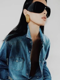 Model wearing oversized black shield sunglasses, a gold statement earring, and an unbuttoned blue denim shirt.