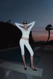 Model Valery Kaufman wearing a white wrap top, capri leggings, sunglasses, and black heels by an outdoor pool at dusk.