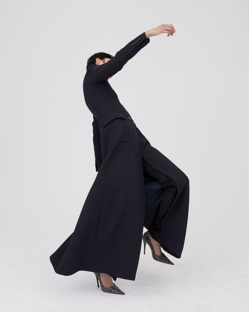 Fashion model in dynamic motion wearing an elongated floor-length dark navy tailored coat and pointed stiletto heels on a white seamless backdrop.