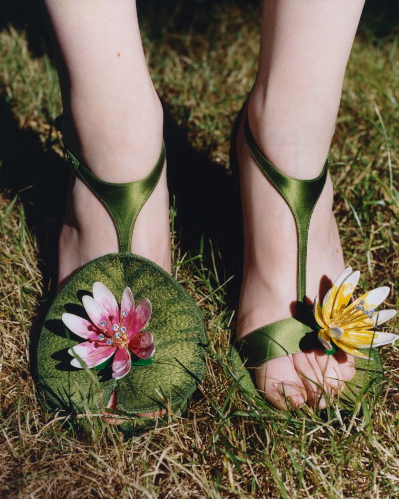 Close-up of feet wearing green satin T-strap sandals adorned with sculptural water lily flowers standing on natural grass.