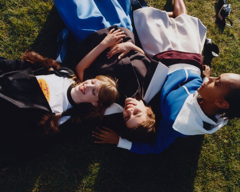 Three female models styled in vibrant outerwear lying closely together on green grass under bright sunlight for WSJ Magazine.