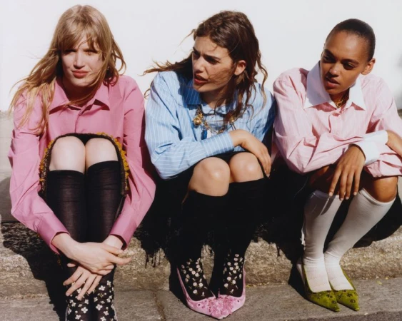 Three models wearing button-down collared shirts, knee-high socks, and pointed-toe heels sitting together against a white wall.