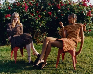 Two fashion models seated on red chairs in front of a rose bush on a green lawn.