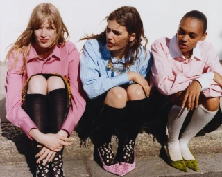 Three models wearing button-down collared shirts, knee-high socks, and pointed-toe heels sitting together against a white wall.