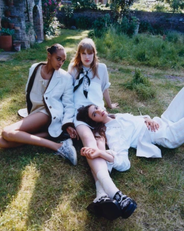 Three female models in tailored white outfits relaxing together on a sunlit grassy lawn in a lush garden.
