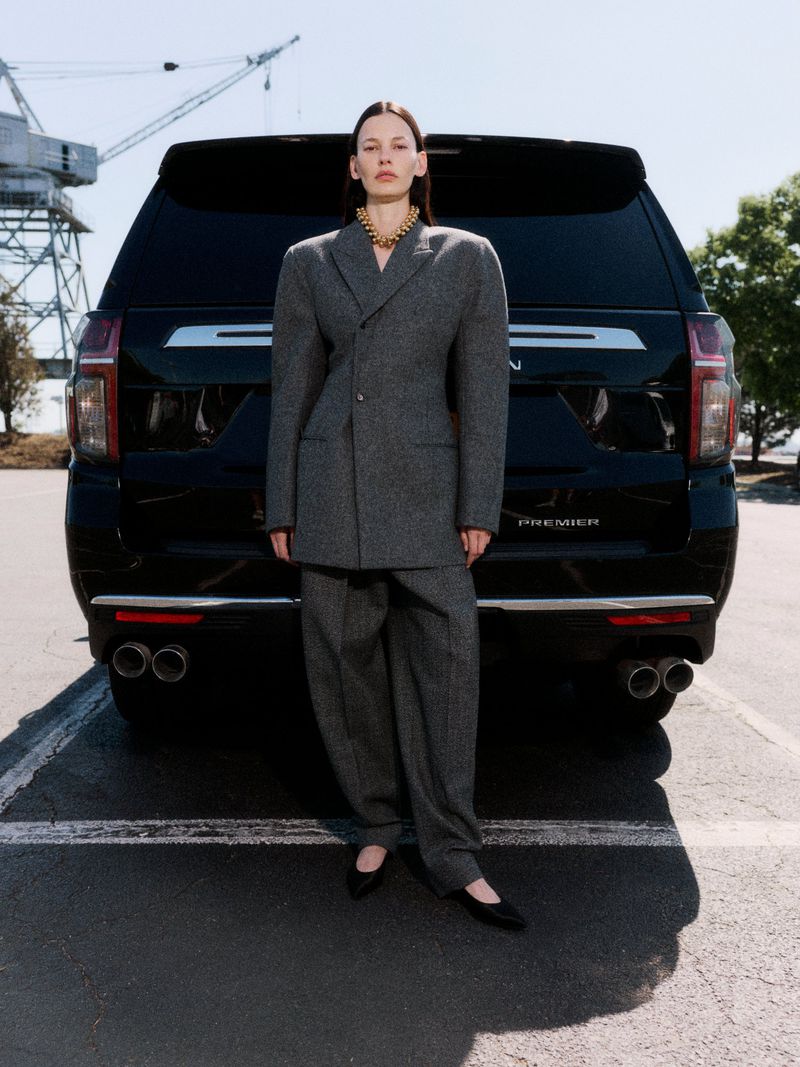 Model Amanda Murphy in an oversized grey wool suit and gold necklace standing in front of a black SUV in an open parking lot.