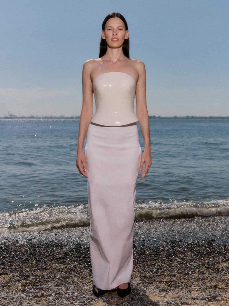Model Amanda Murphy stands on a pebble beach wearing a strapless glossy cream corset top and a long white column skirt.