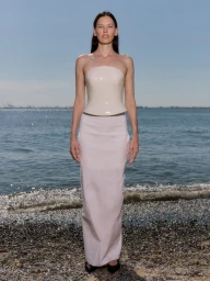 Model Amanda Murphy stands on a pebble beach wearing a strapless glossy cream corset top and a long white column skirt.
