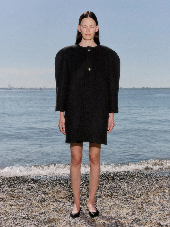 Model Amanda Murphy standing on a pebbled beach wearing a structured black round-shoulder coat dress and black flats.