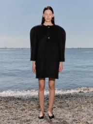 Model Amanda Murphy standing on a pebbled beach wearing a structured black round-shoulder coat dress and black flats.