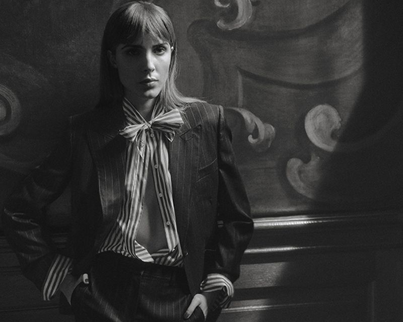 Black and white portrait of a woman wearing a pinstripe suit and striped bow blouse standing against a dark decorative panelled wall.