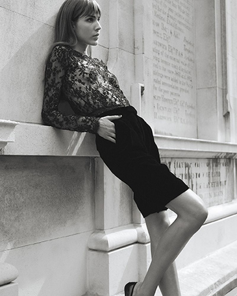 Model Freya Aspinall leaning against an inscribed stone wall wearing a sheer black lace long-sleeve top and tailored black shorts.